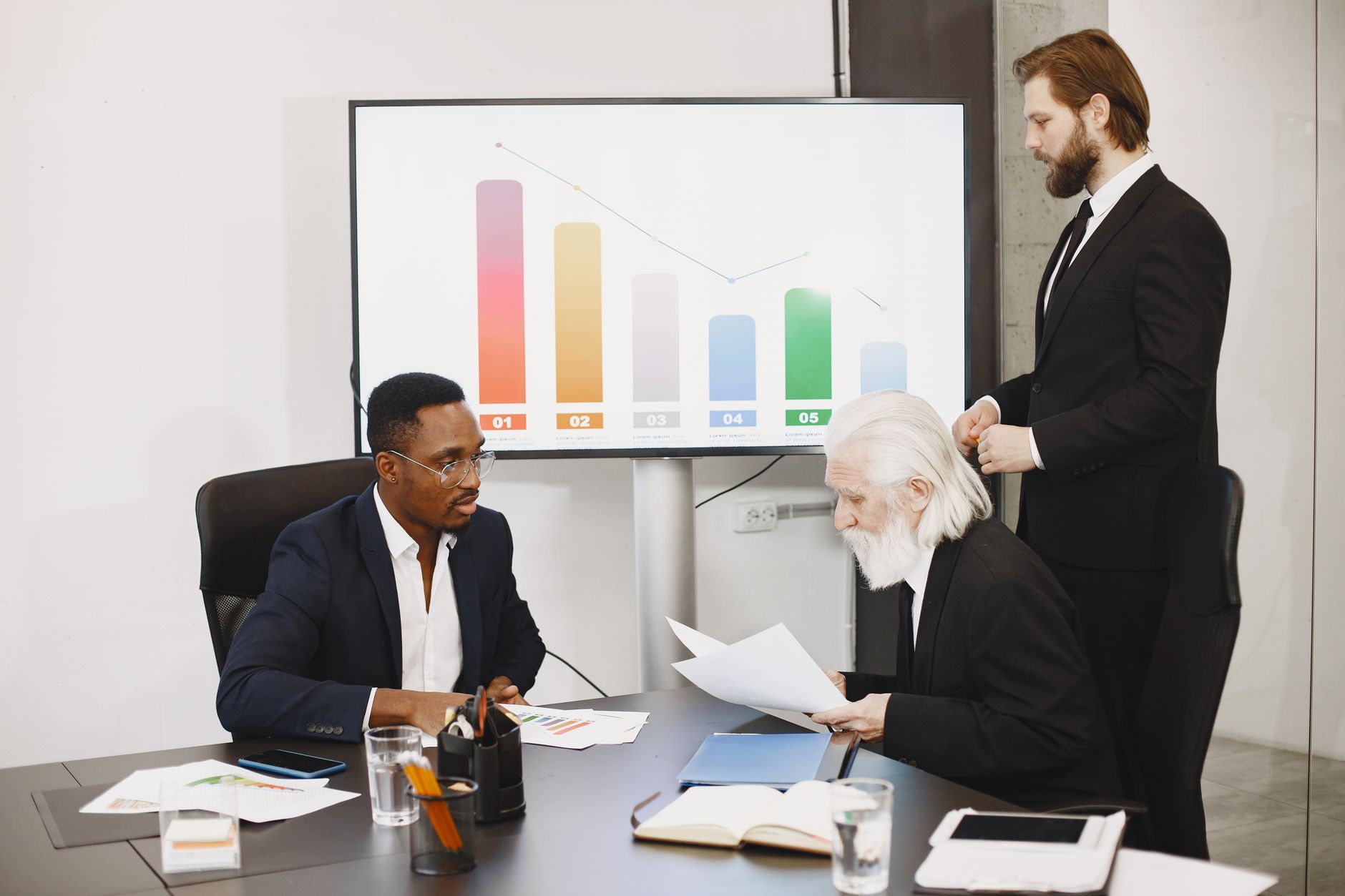 Three businessmen discussing financial data in a modern office setting with charts. — Brody Shulman Providing Comprehensive R