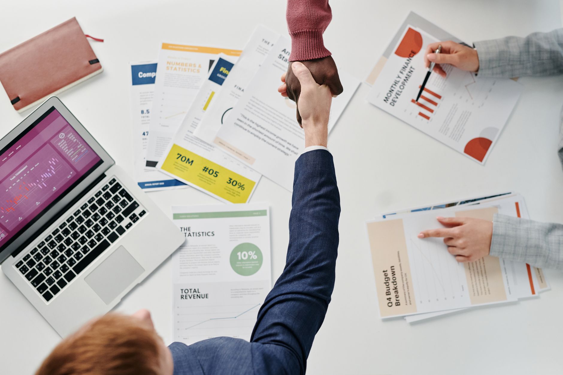 Business professionals engaging in a handshake over a desk filled with financial documents and a laptop. — Kevin D. Kuykendal