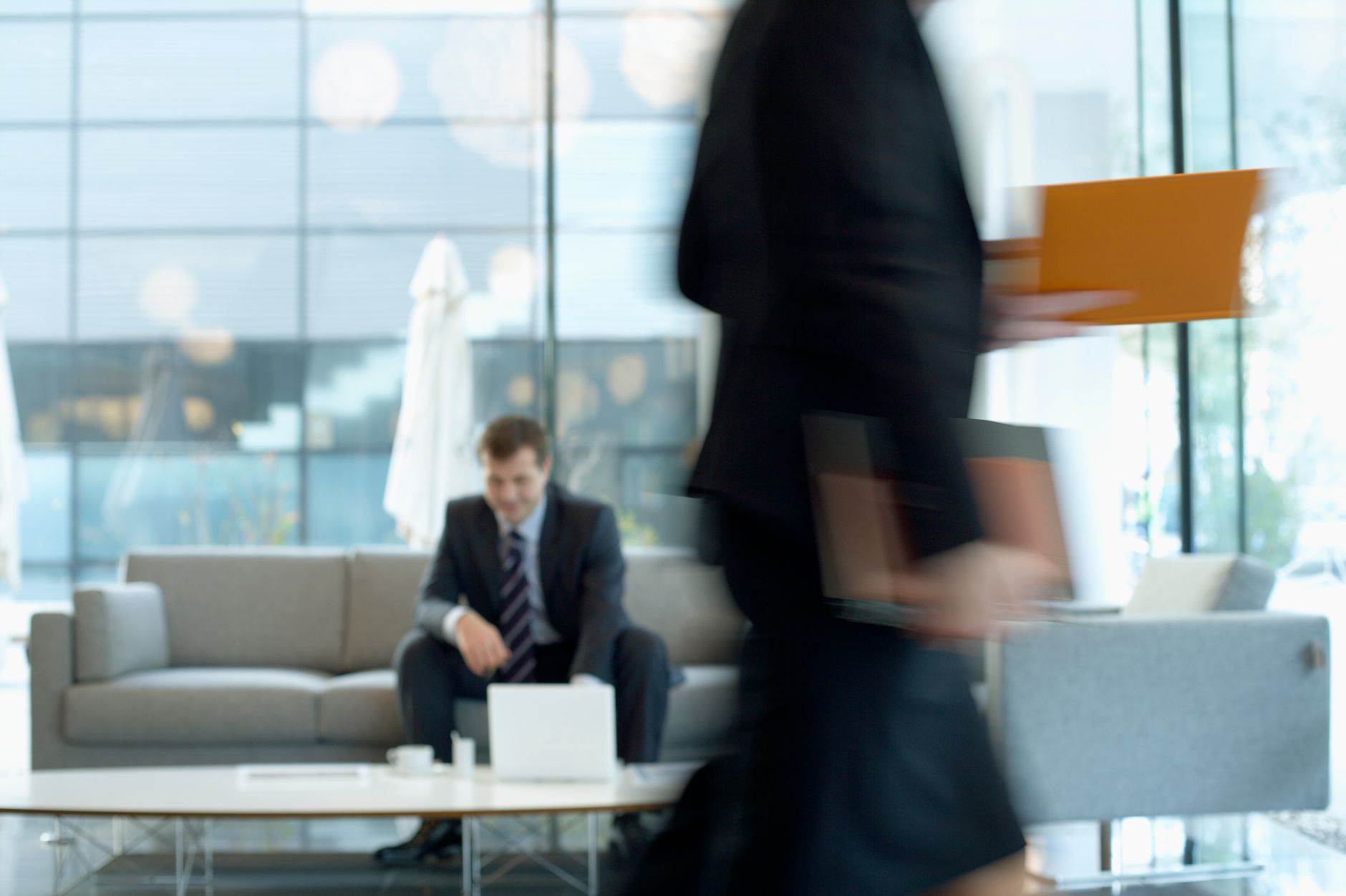Businessman sitting in modern office lounge using a laptop while colleague walks past, creating dynamic blur. — The Evolution