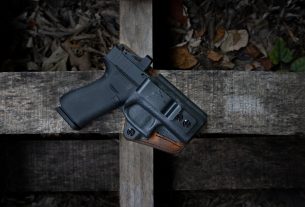 A handgun in a holster resting on wooden planks covered with leaves.